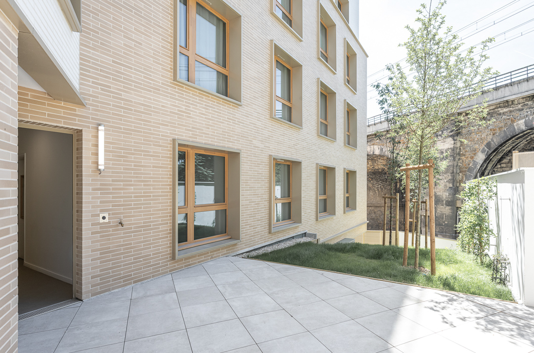Espace extérieur privé d'un centre d'hébergement d'urgence avec façade en plaquettes en terre cuite sable et larges fenêtres en bois